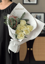 Bouquet of white roses with greenery and a red ribbon, branded 'HANAKO', held by a person.