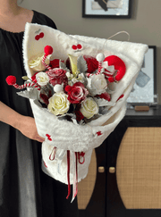 Bouquet of red and white flowers wrapped in white paper with decorative elements, held by a person.