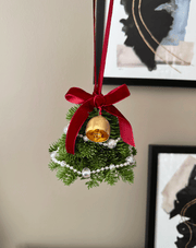 Decorative Christmas ornament with a gold bell, red ribbon, and greenery on a neutral background.