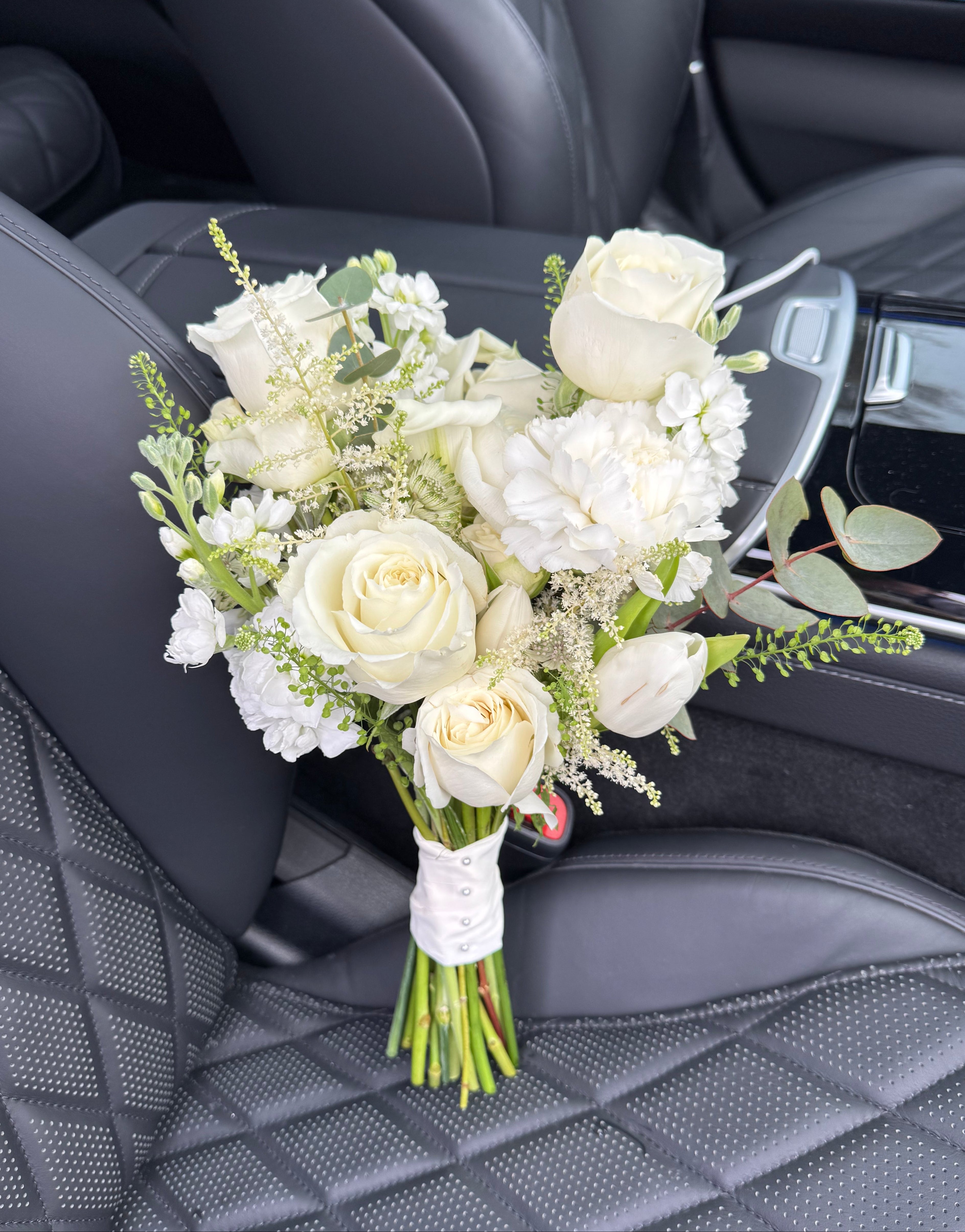 A bridal bouquet of white roses, white carnations, baby's breath, and green foliage, wrapped in a white ribbon and resting on a black quilted leather car seat.