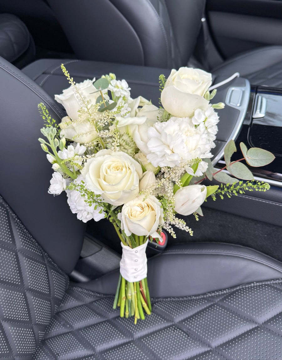 A bridal bouquet of white roses, white carnations, baby's breath, and green foliage, wrapped in a white ribbon and resting on a black quilted leather car seat.